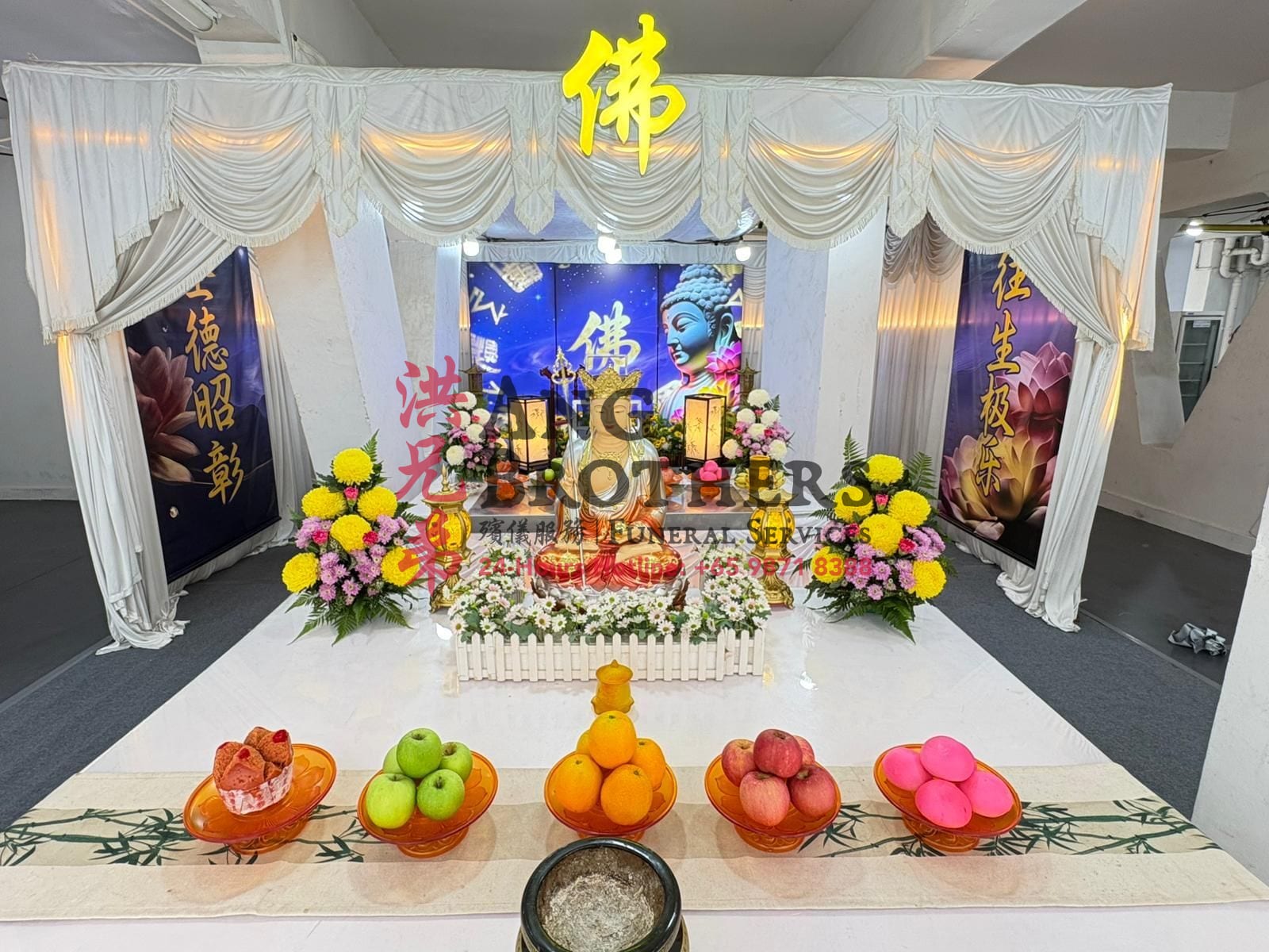 A Buddhist funeral altar with a golden bodhisattva beneath a Buddha backdrop, dressed with flowers and fruit offerings by Ang Brothers